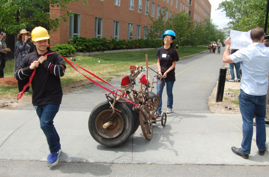 “Apoplectic Apocalyptics” was another of Bill Turville’s contraptions, in this case built from “trash recovered from the Lowell canal system” during a clean-up he took part in some years back. “The wheel is made out of wood and it’s cracked,” said Amy Xiao (center), a River Fest volunteer who was roped into racing the thing. “So we had to drag it.” (Greg Cook)